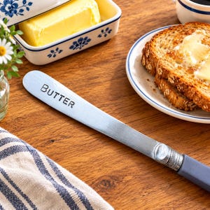 Op de afbeelding: Een zilverkleurig metalen botermes met een glad, plat lemmet waarop het woord BUTTER in zwarte hoofdletters is gestempeld. Het handvat heeft een effen, matte grijze kleur en het keukengerei heeft een lang, slank profiel om mee te smeren.
