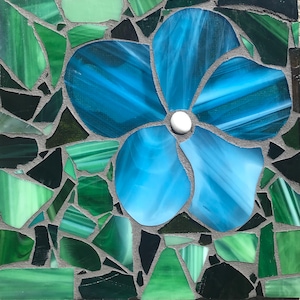 May include: A blue flower mosaic made from small pieces of glass, set against a background of green and black glass pieces.