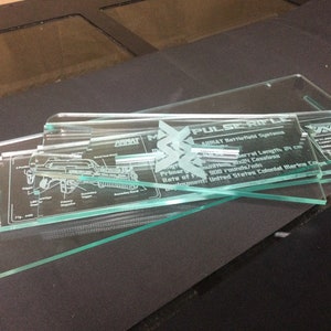以下が含まれることがあります： レーザーエッチングされた透明ガラスのディスプレイピース。「PULSE RIFLE」の文字、技術仕様、図面が見えます。デザインにはARMATのロゴやその他のテキストが含まれています。これらのピースは暗い表面に積み重ねられています。