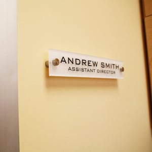 May include: A white rectangular office door sign with black text that reads "ANDREW SMITH ASSISTANT DIRECTOR".