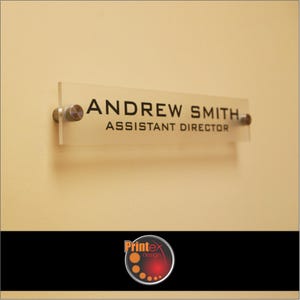 May include: A clear acrylic office nameplate with the name "ANDREW SMITH" and title "ASSISTANT DIRECTOR" in black lettering. The nameplate is mounted on a beige wall with silver-colored standoffs. The Printex design logo is at the bottom.