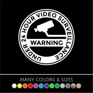 May include: A black and white circular sign with the text "UNDER 24 HOUR VIDEO SURVEILLANCE WARNING". The sign features a security camera icon. The bottom of the sign says "MANY COLORS & SIZES" with color options.