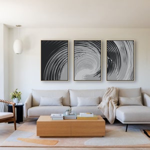 May include: Three abstract black and white swirl paintings in gold frames hanging on a white wall above a gray sectional sofa with a throw blanket and a wooden coffee table.