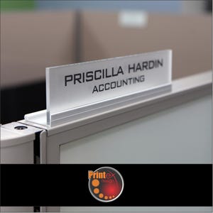 May include: A clear acrylic desk nameplate with the name "PRISCILLA HARDIN" and the title "ACCOUNTING" in black lettering. The nameplate is mounted on a silver metal frame, designed for office cubicles. The Printex design logo is at the bottom.