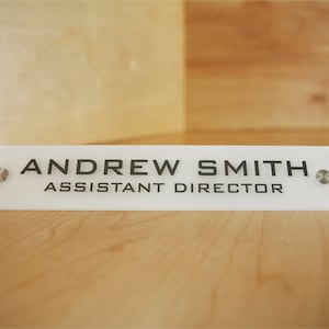 May include: White acrylic desk nameplate with black text that reads "ANDREW SMITH ASSISTANT DIRECTOR". The nameplate has two silver screws on either side.