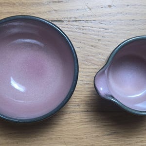 May include: A circular ceramic bowl and a matching small ceramic pitcher with a pouring spout. Both items feature a smooth, matte mauve interior and a dark, charcoal-colored rim, showcasing a consistent glaze finish across both pieces.