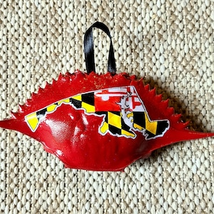 May include: A red crab-shaped ornament with a black ribbon for hanging. The ornament features the Maryland state flag design in the center. The crab has a textured, spiky edge.