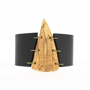May include: A black leather cuff bracelet with a triangular wooden accent. The wood has a natural grain pattern and is secured to the leather with brass rivets.