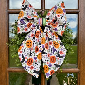 May include: A large decorative bow featuring a white fabric with a repeating Halloween pattern of orange pumpkins, white ghosts, black cats, bats, skulls wearing purple hats, and small purple and red flowers, finished with a striped center knot.