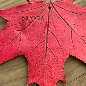 K&ouml;nnte beinhalten: Ein rotes Ahornblatt mit dem eingepr&auml;gten Wort "CANADA". Das Blatt ist aus einem strukturierten Material gefertigt und hat oben ein Loch zum Aufh&auml;ngen.