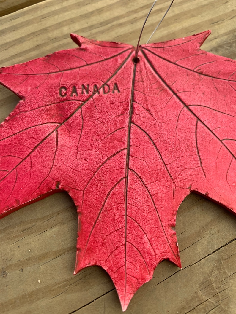 K&ouml;nnte beinhalten: Ein rotes Ahornblatt mit dem eingepr&auml;gten Wort "CANADA". Das Blatt ist aus einem strukturierten Material gefertigt und hat oben ein Loch zum Aufh&auml;ngen.