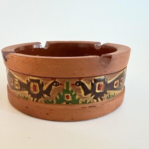 May include: A round terracotta ashtray featuring a matte finish and a decorative band around the exterior. The band displays a repeating pattern of stylized dark brown birds with red and gold accents, separated by green geometric tree-like shapes.