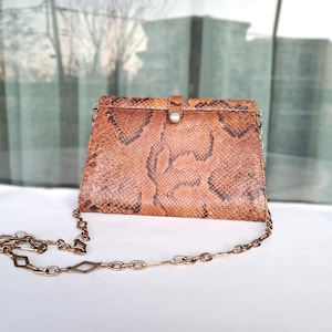 Puede incluir: Bolso de mano rectangular de piel de serpiente marrón con cadena dorada. El bolso tiene un cierre de solapa con un botón plateado. El estampado de piel de serpiente incluye marcas marrones oscuras y negras. La cadena es una mezcla de eslabones redondos y en forma de diamante.