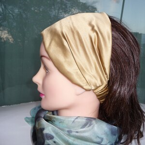 May include: A gold satin headband worn on a mannequin with brown hair. The headband is folded over and tied at the back of the head.