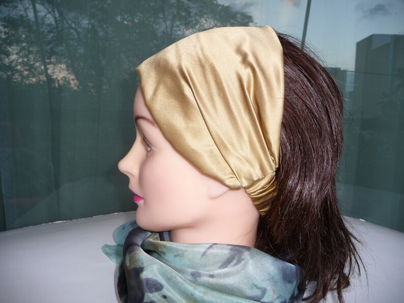 May include: A gold satin headband worn on a mannequin with brown hair. The headband is folded over and tied at the back of the head.