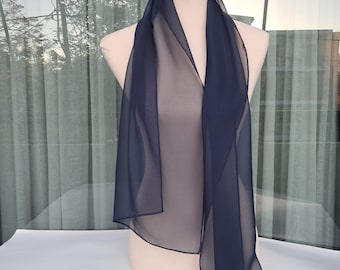 May include: A navy blue scarf displayed on a mannequin. The scarf is made of a sheer, lightweight material. The scarf is long and rectangular, with a simple, elegant design. The background includes a window with a view of trees and a white surface.