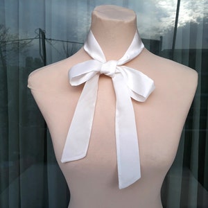 May include: A white satin bow tie on a white mannequin. The bow tie is tied in a simple knot and the ends are hanging down.