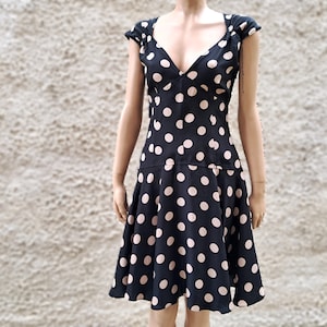 Könnte beinhalten: Ein schwarzes Kleid mit V-Ausschnitt und Flügelärmeln, verziert mit großen, gleichmäßig verteilten rosa Punkten. Das Kleid hat ein figurbetontes Oberteil und einen ausgestellten Rock, wodurch eine klassische Silhouette entsteht. Der Stoff scheint leicht zu sein.