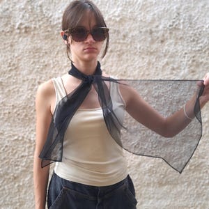 May include: A woman wearing a black sheer scarf tied around her neck. She is wearing a white tank top and black jeans. She is also wearing sunglasses.