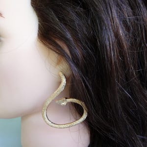 Peut inclure: Une boucle d'oreille dorée en forme de serpent enroulé avec une surface texturée rappelant des écailles. La boucle d'oreille se courbe en une forme circulaire, la tête du serpent pointant vers l'intérieur du cercle, créant un design ouvert.