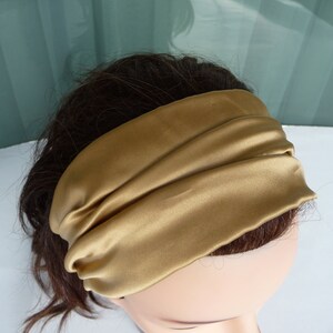 May include: A gold satin headband with a simple, folded design. The headband is worn on a person's head.