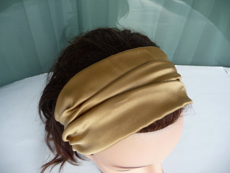 May include: A gold satin headband with a simple, folded design. The headband is worn on a person's head.
