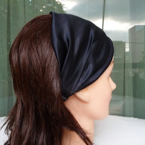 May include: A black satin headband worn on a person's head. The headband is folded over and has a smooth, silky texture.