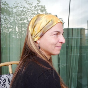 May include: A woman with long blonde hair is wearing a yellow and brown patterned headband. She is looking to the right and smiling.