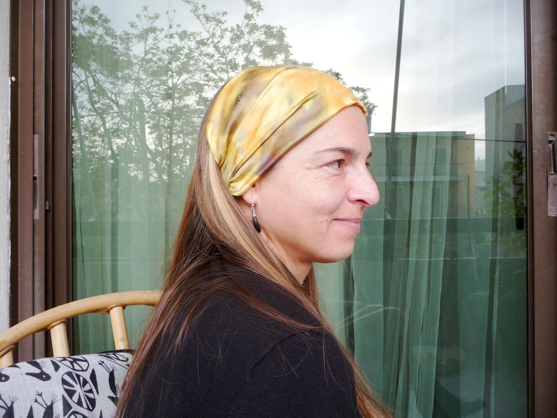May include: A woman with long blonde hair is wearing a yellow and brown patterned headband. She is looking to the right and smiling.