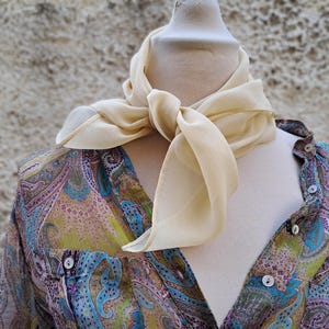 May include: A cream-coloured scarf is knotted around the neck of a mannequin. The scarf is draped over a button-down shirt with a colourful paisley pattern in shades of blue, green, and purple.
