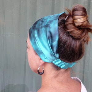 May include: A teal blue satin headband with a tie-dye pattern. The headband is worn around the head and tied at the back of the neck.