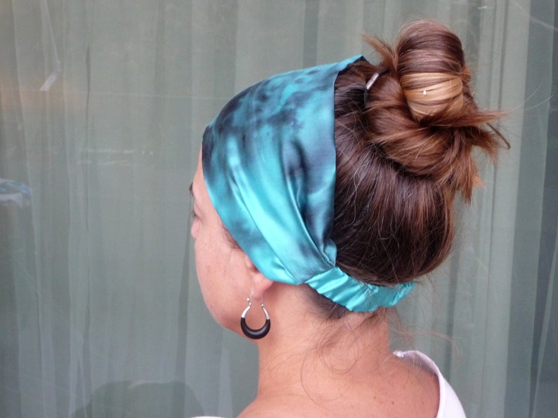 May include: A teal blue satin headband with a tie-dye pattern. The headband is worn around the head and tied at the back of the neck.