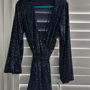 May include: A black sequined mini dress with long sleeves and a V-neckline. The dress has a tie belt at the waist and a flowing skirt.