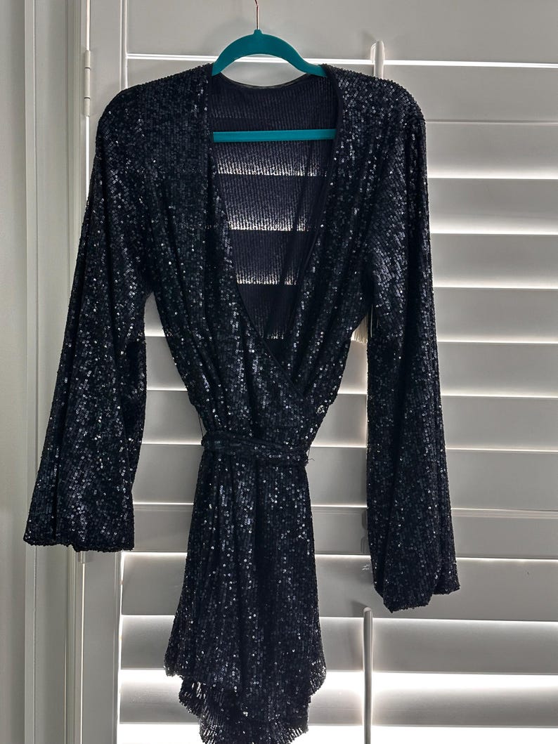 May include: A black sequined mini dress with long sleeves and a V-neckline. The dress has a tie belt at the waist and a flowing skirt.