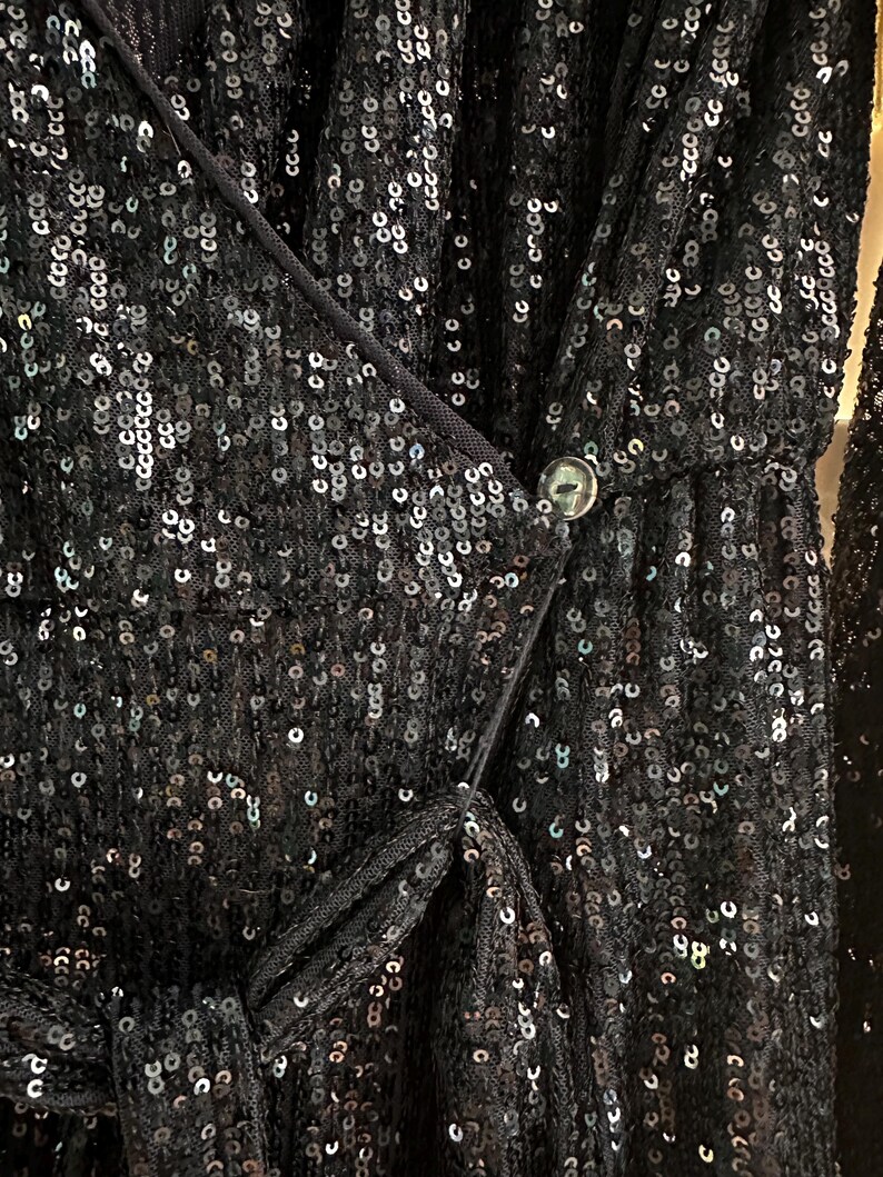 May include: A black sequined dress with a wrap-style bodice and a button closure. The dress is made of a sheer fabric with sequins that shimmer in the light.