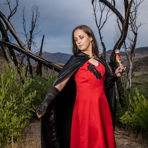 May include: A woman in a red dress and black cape holds a glass of red liquid. She is standing in a forest setting with dead trees and a skull on the ground.