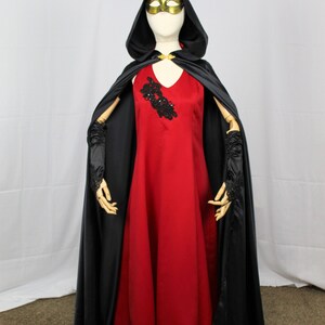 May include: A red dress with a black lace embellishment on the bodice, worn with a black hooded cape and matching gloves. The model is wearing a gold mask.