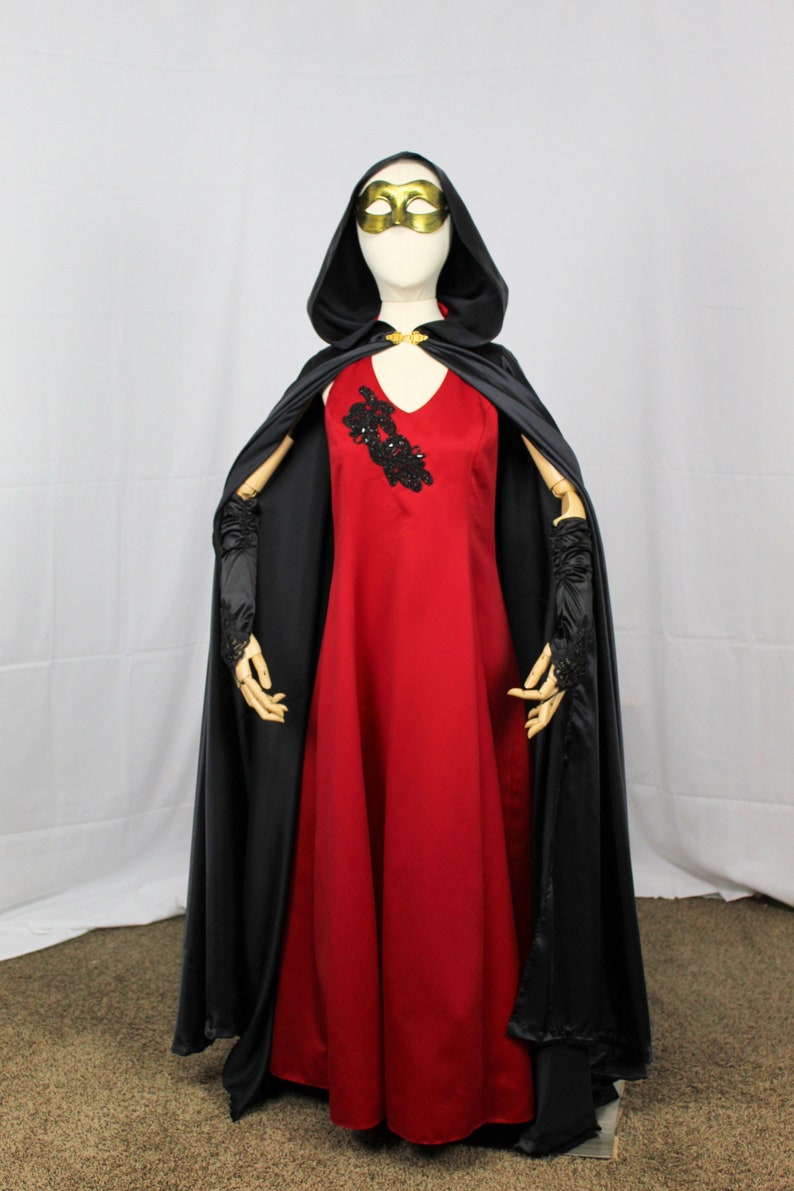 May include: A red dress with a black lace embellishment on the bodice, worn with a black hooded cape and matching gloves. The model is wearing a gold mask.