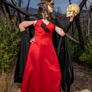 May include: A woman in a red dress and black cape holds a human skull in her right hand. She is wearing a black mask over her eyes. The background is a blurred image of a forest.