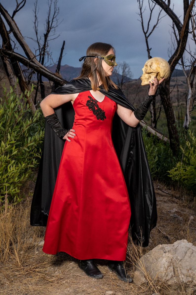 May include: A woman in a red dress and black cape holds a human skull in her right hand. She is wearing a black mask over her eyes. The background is a blurred image of a forest.