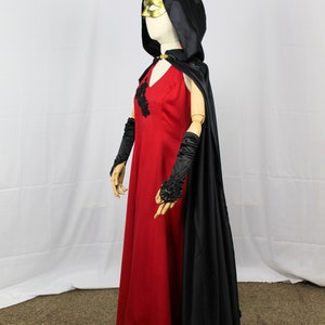 May include: A red dress with black lace detailing and a black satin cape with a hood. The dress has a v-neckline and the cape has a gold mask attached to the hood.