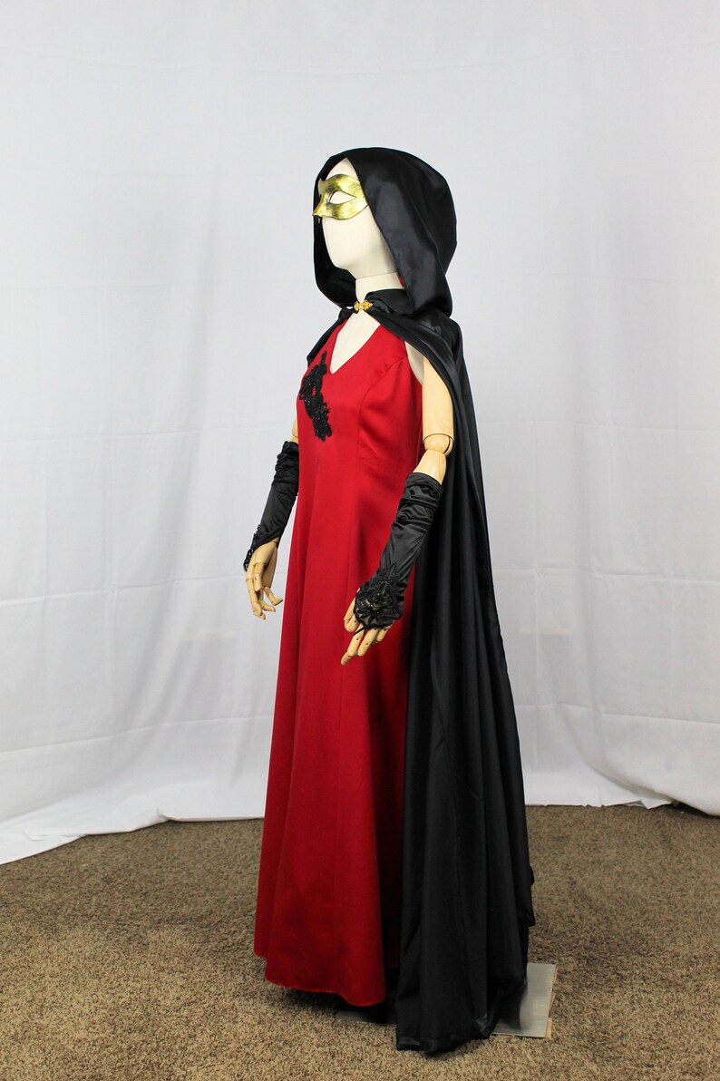 May include: A red dress with black lace detailing and a black satin cape with a hood. The dress has a v-neckline and the cape has a gold mask attached to the hood.