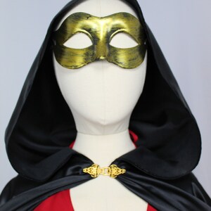 May include: A black satin cape with a gold clasp and a gold masquerade mask. The cape has a hood and is worn over a red dress.