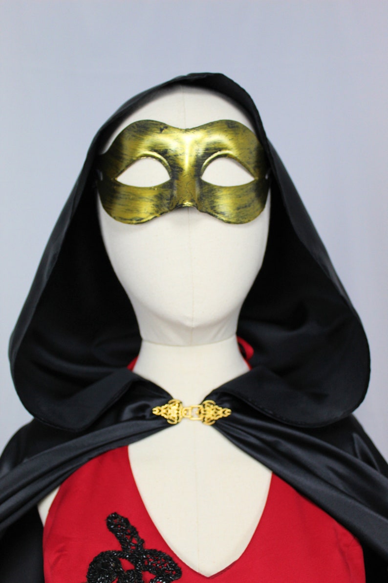 May include: A black satin cape with a gold clasp and a gold masquerade mask. The cape has a hood and is worn over a red dress.