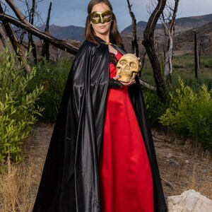 May include: A woman wearing a black cape and a red dress holds a white skull in her hands. She is wearing a gold mask over her eyes. The background is a forest with dead trees.