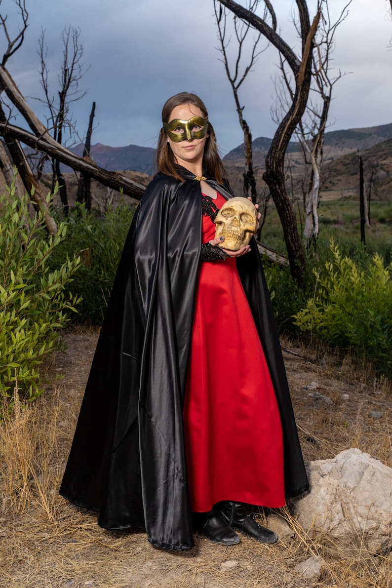 May include: A woman wearing a black cape and a red dress holds a white skull in her hands. She is wearing a gold mask over her eyes. The background is a forest with dead trees.