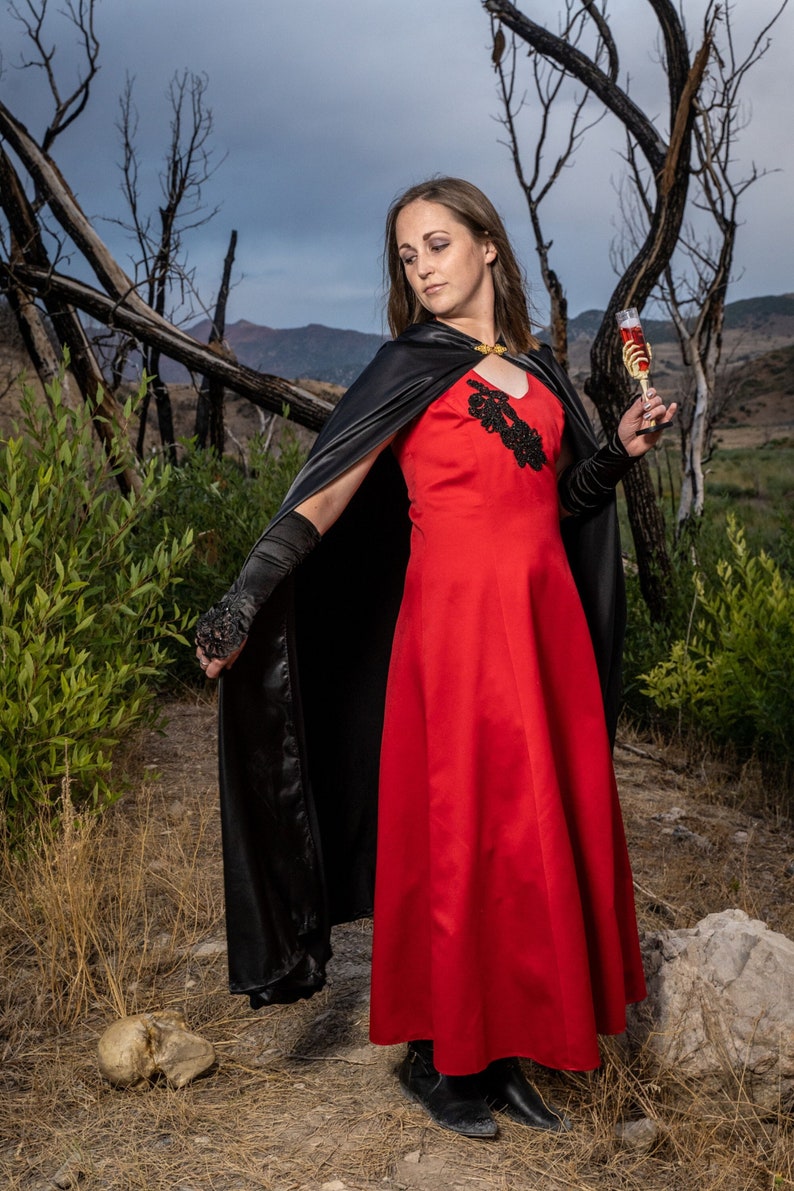 May include: A woman in a red dress and black cape holds a glass of red liquid. She is standing in a forest setting with dead trees and a skull on the ground.
