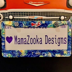 May include: A white license plate frame with a blue and yellow starry night design. The frame has a purple heart and the text "MamaZoooka Designs" in purple. The frame also has the text "Est 1997" and "Handmade".