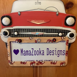 May include: A red, white, and black vintage car-shaped license plate frame with a pink and white floral pattern. The frame has a purple heart with the text "MamaZoooka Designs" and the text "License Plate Frames".