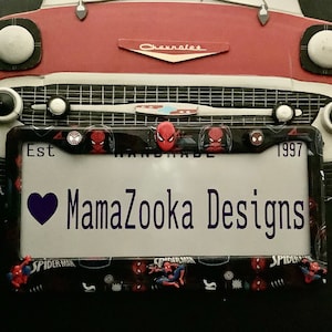 May include: A black and red license plate frame with a Spider-Man theme. The frame has a heart with the text "MamaZoooka Designs" and the years "Est 1997".